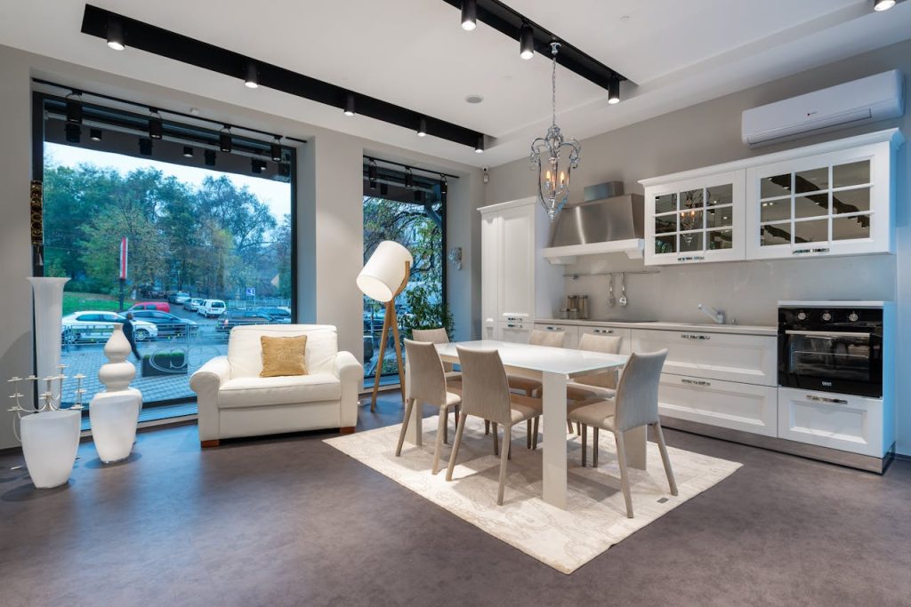 Modern open-plan kitchen and dining area with white cabinetry, glass-front upper cabinets, stainless steel appliances, white dining table with six beige chairs, large windows, white armchair with gold cushion