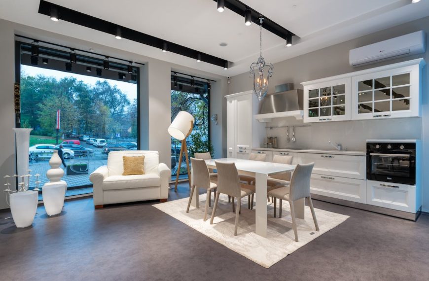 Modern open-plan kitchen and dining area with white cabinetry, glass-front upper cabinets, stainless steel appliances, white dining table with six beige chairs, large windows, white armchair with gold cushion