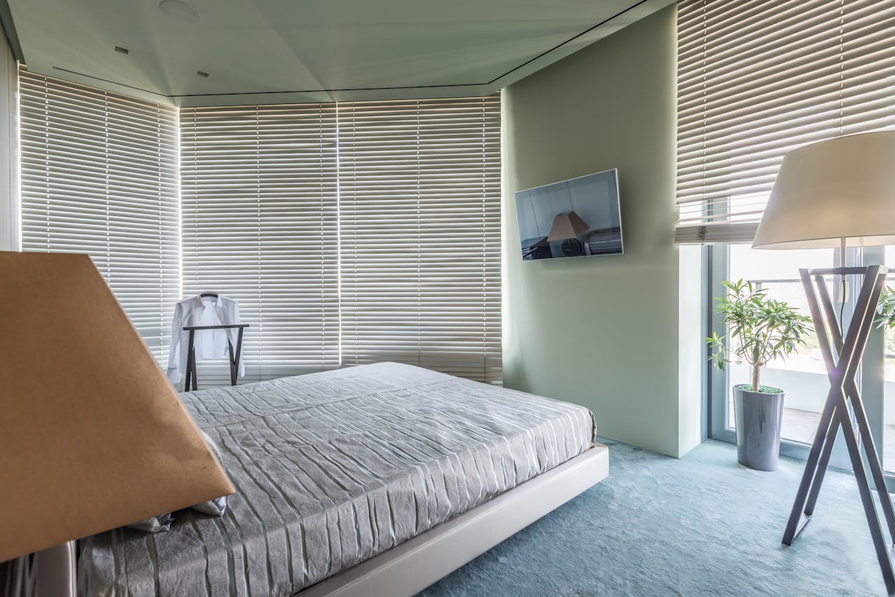 Modern minimalist bedroom, floor-to-ceiling blinds, wall-mounted TV, floor lamp, potted plant, neatly made bed with gray bedding, light green walls, soft blue carpet, shirt draped on chair