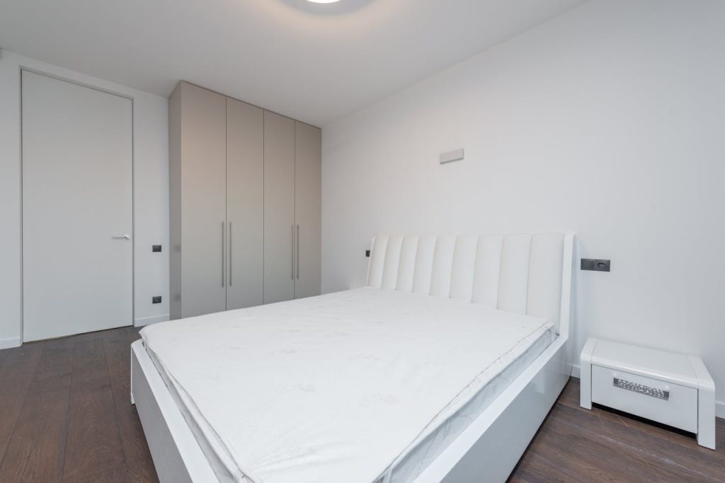 Modern minimalist bedroom, white padded headboard bed frame, white mattress, white bedside table, beige built-in wardrobes, dark wooden floor, white walls
