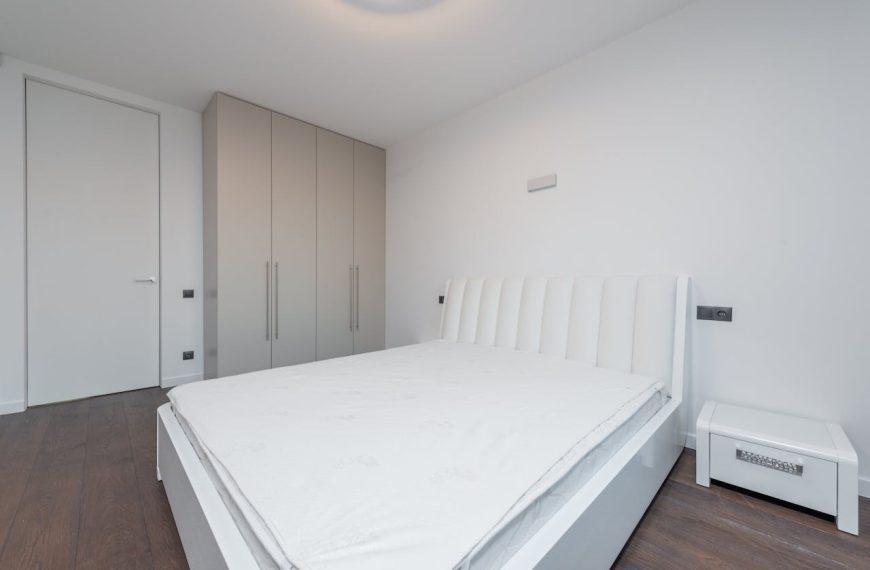 Modern minimalist bedroom, white padded headboard bed frame, white mattress, white bedside table, beige built-in wardrobes, dark wooden floor, white walls