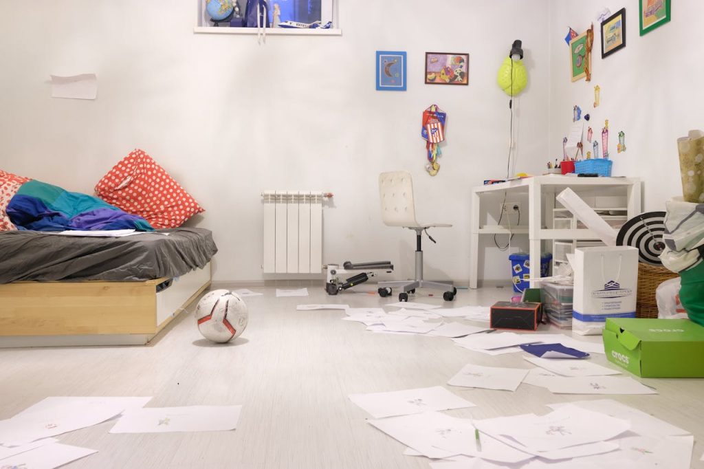 Messy children's bedroom, unmade bed with red pillows, papers scattered on the floor, soccer ball, white desk and chair, toys and school supplies, colorful wall decorations
