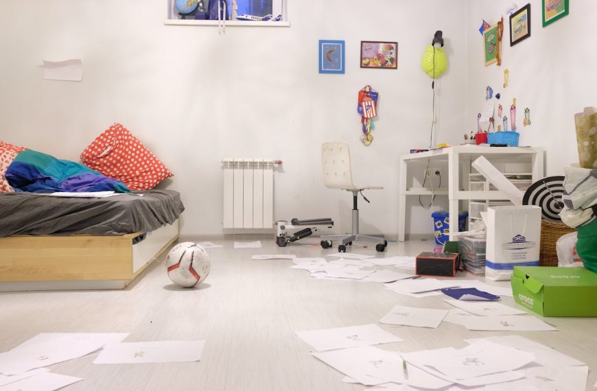 Messy children's bedroom, unmade bed with red pillows, papers scattered on the floor, soccer ball, white desk and chair, toys and school supplies, colorful wall decorations