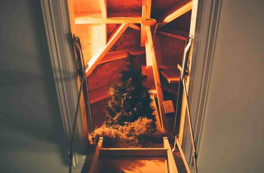 Dimly lit attic with exposed wooden beams, a folded ladder leading up, artificial Christmas tree and decorations stored inside