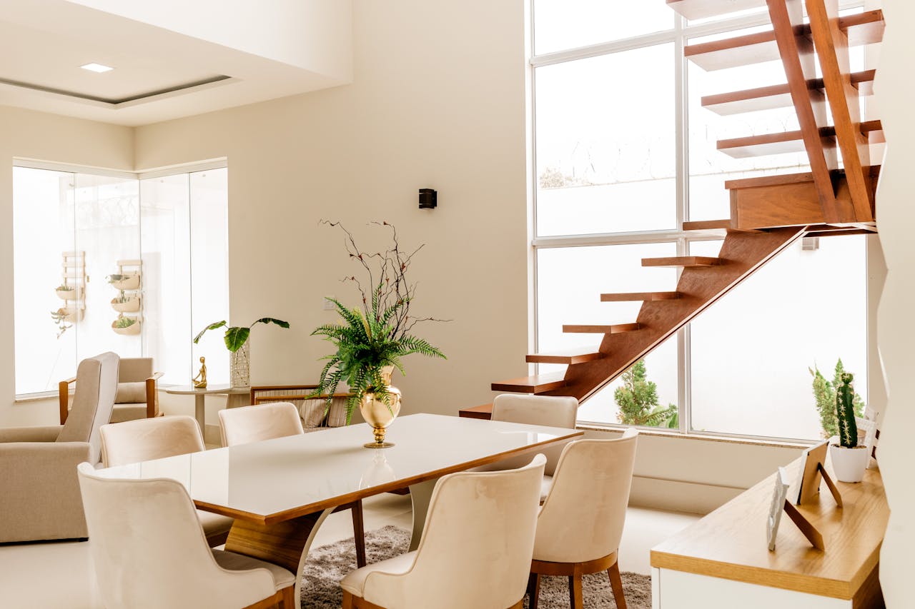 Modern dining room with a wooden staircase, beige chairs around a white table, a gold vase with green plants, large windows with natural light, and neutral-toned decor