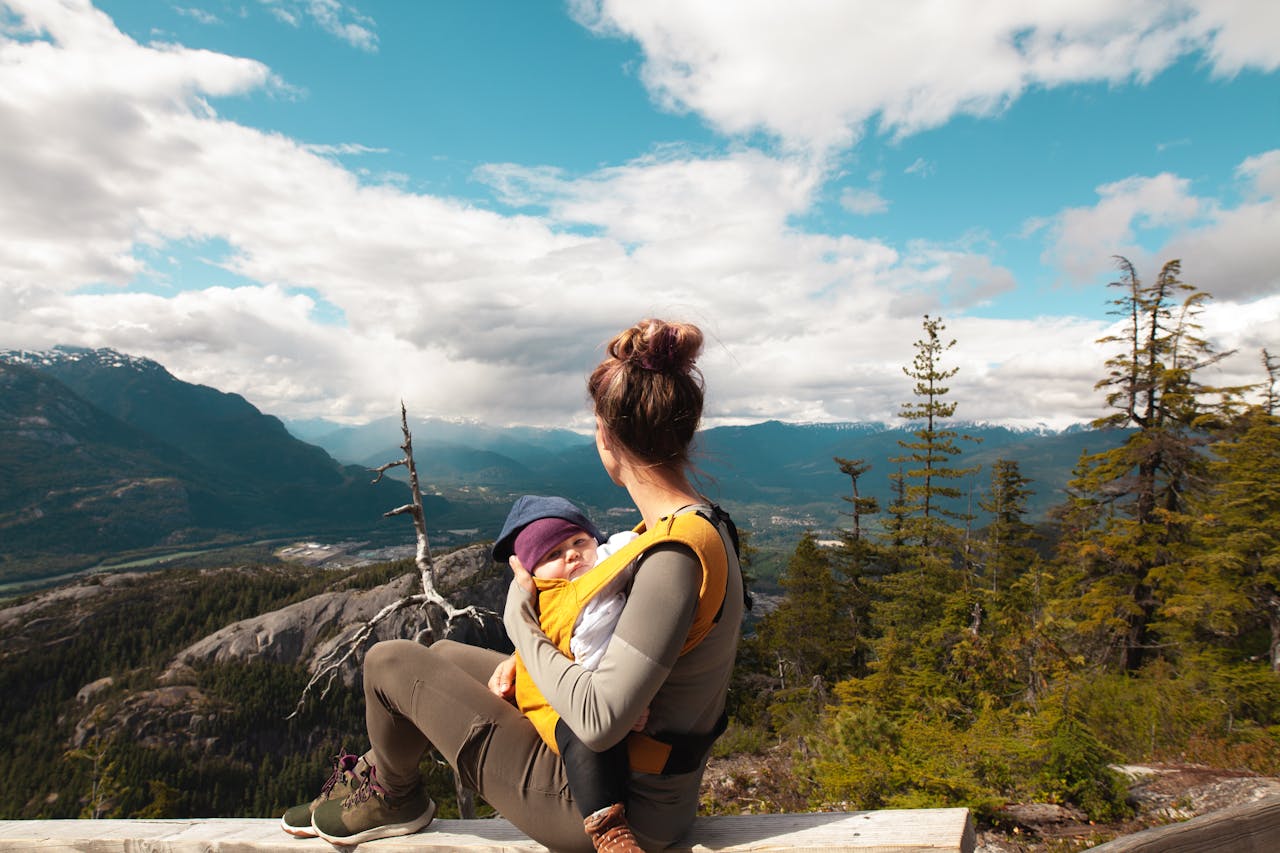 Woman sitting on a wooden bench, holding a baby in a front carrier, facing a scenic mountain landscape under a partly cloudy sky, surrounded by trees