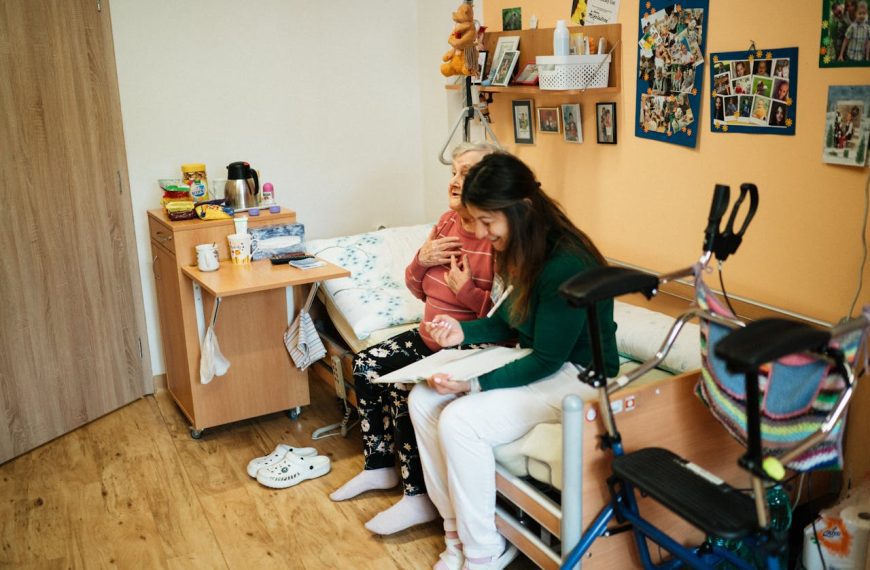 Elderly woman sitting on a bed beside a younger female caregiver, both smiling and laughing, cozy room with wooden furniture, medical walker in foreground,