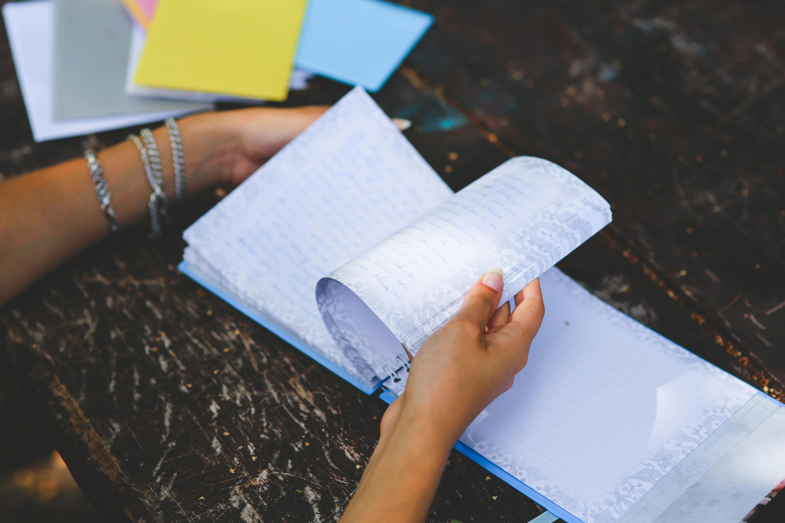 Hands flipping through a handwritten notebook with decorative borders, open on a worn wooden surface, colorful folders scattered in the background, person wearing silver bracelets, natural outdoor lighting suggests a casual study or journaling session