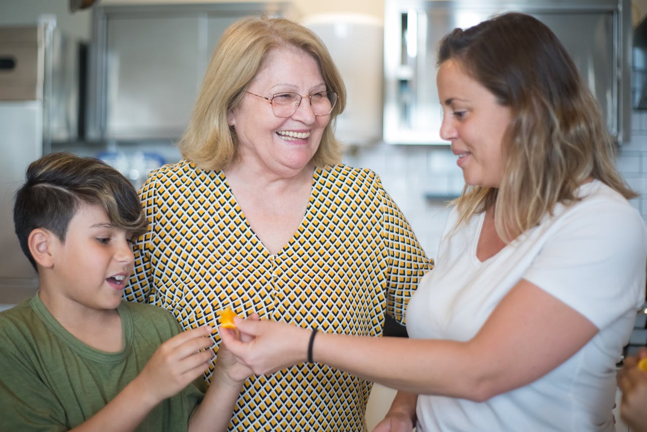 Smiling older woman standing in a kitchen with a younger woman and a boy, all sharing a cheerful moment and passing a piece of fruit