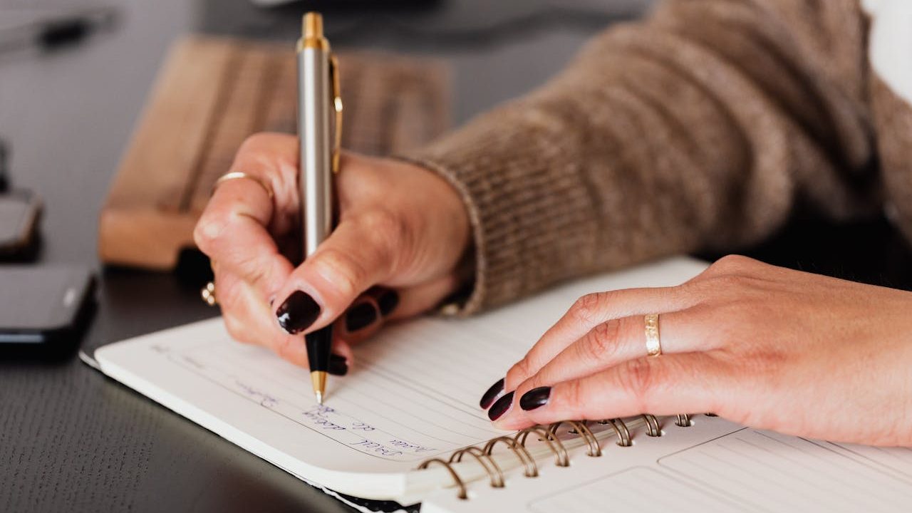Close-up of a woman’s hands writing with a pen in a spiral notebook during a journaling or planning session