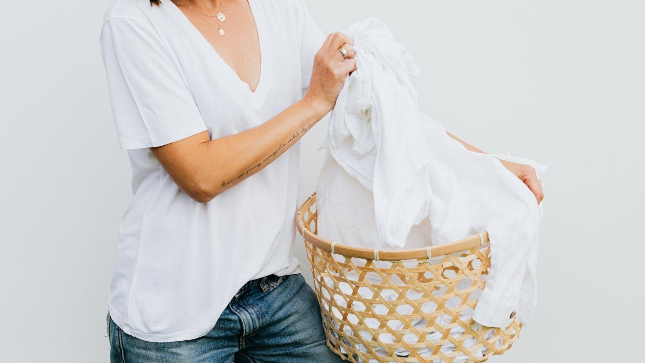 Woman in white t-shirt and jeans holding a woven laundry basket filled with white clothes, mid-action, against a plain white background
