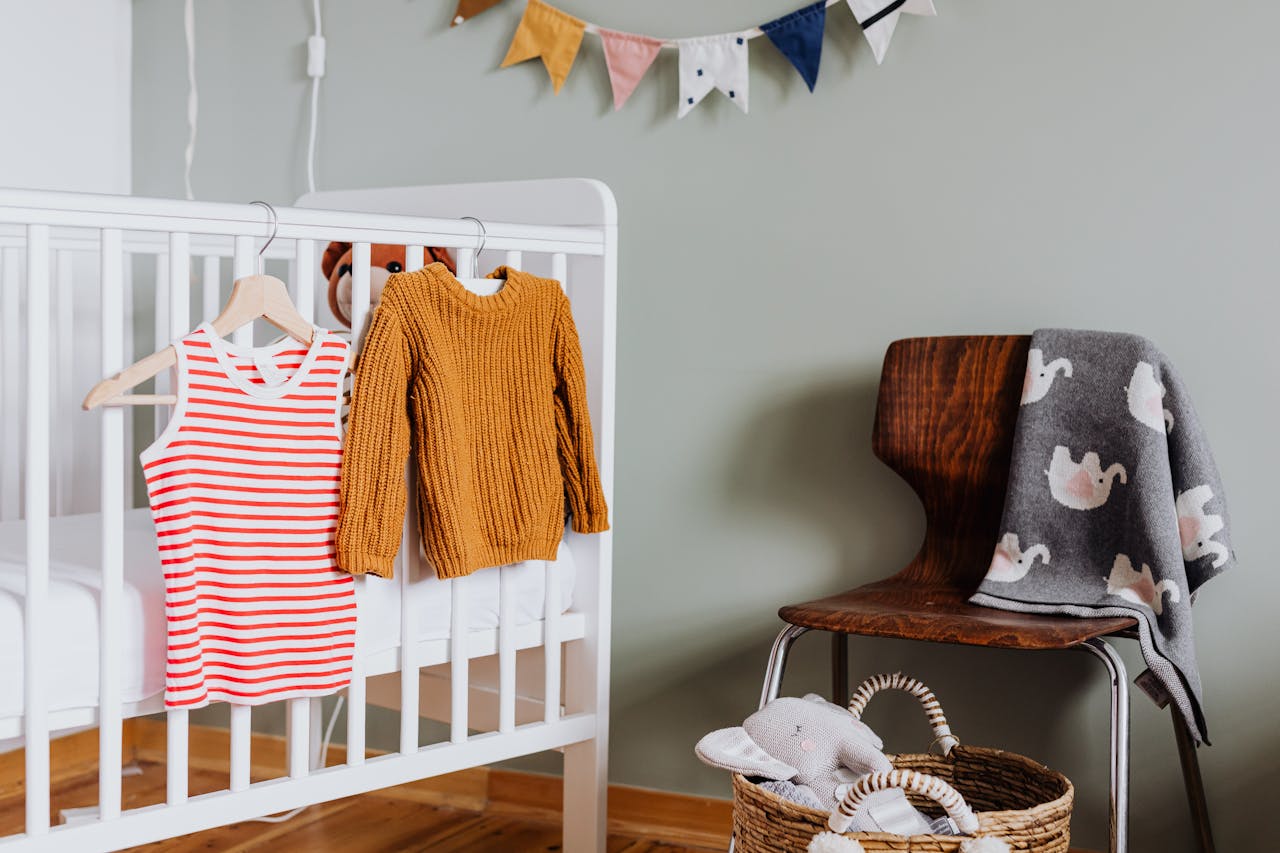 Children’s clothing hanging on a white crib, mustard knit sweater, red and white striped tank top, wooden chair with elephant-print blanket, basket with stuffed animal, colorful bunting on wall