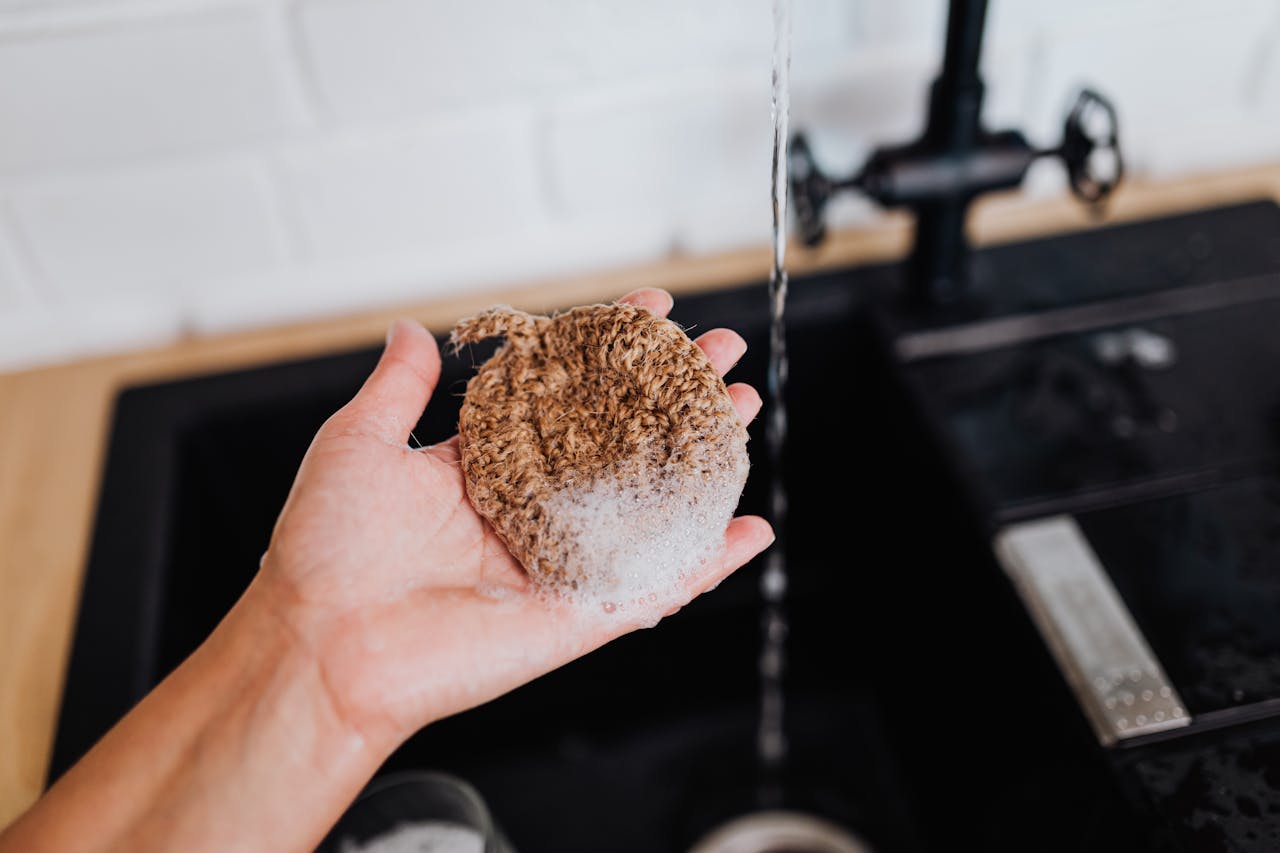 Hand holding a soapy, brown, crocheted scrubber under running water at a black kitchen sink, with white tiled backsplash and wooden countertop