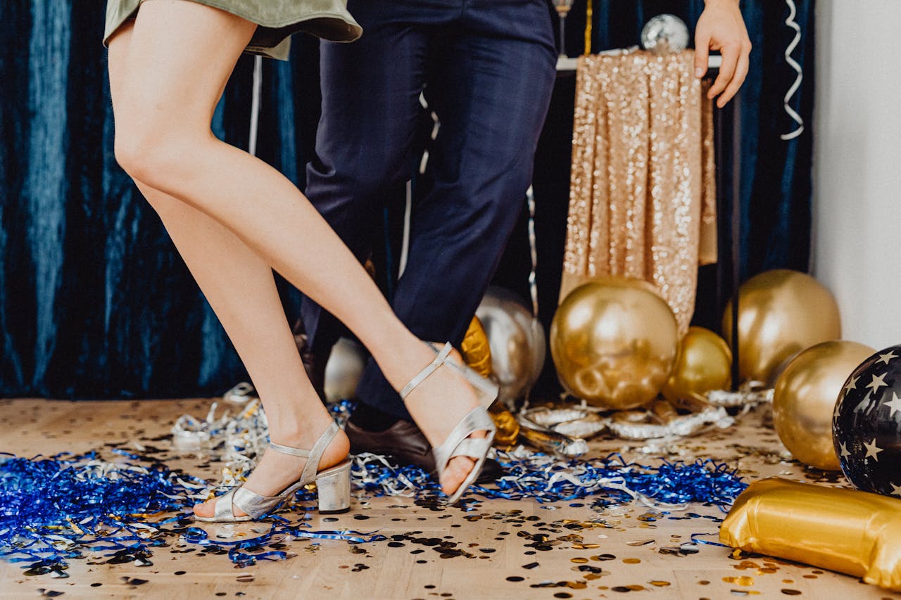 Two people dancing on a confetti-covered floor, woman in silver heels and green dress, man in dark trousers and brown shoes, surrounded by gold and black balloons, blue and silver streamers, festive party setting with a glittery backdrop