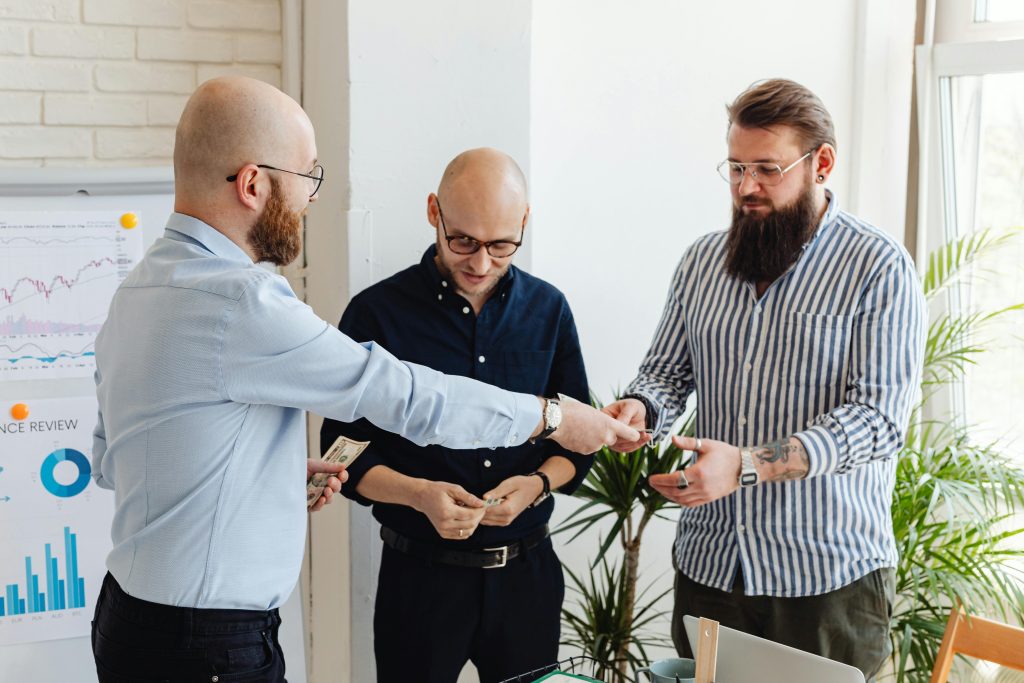 Three men standing in an office space exchanging U.S. dollar bills, with business charts on a whiteboard in the background