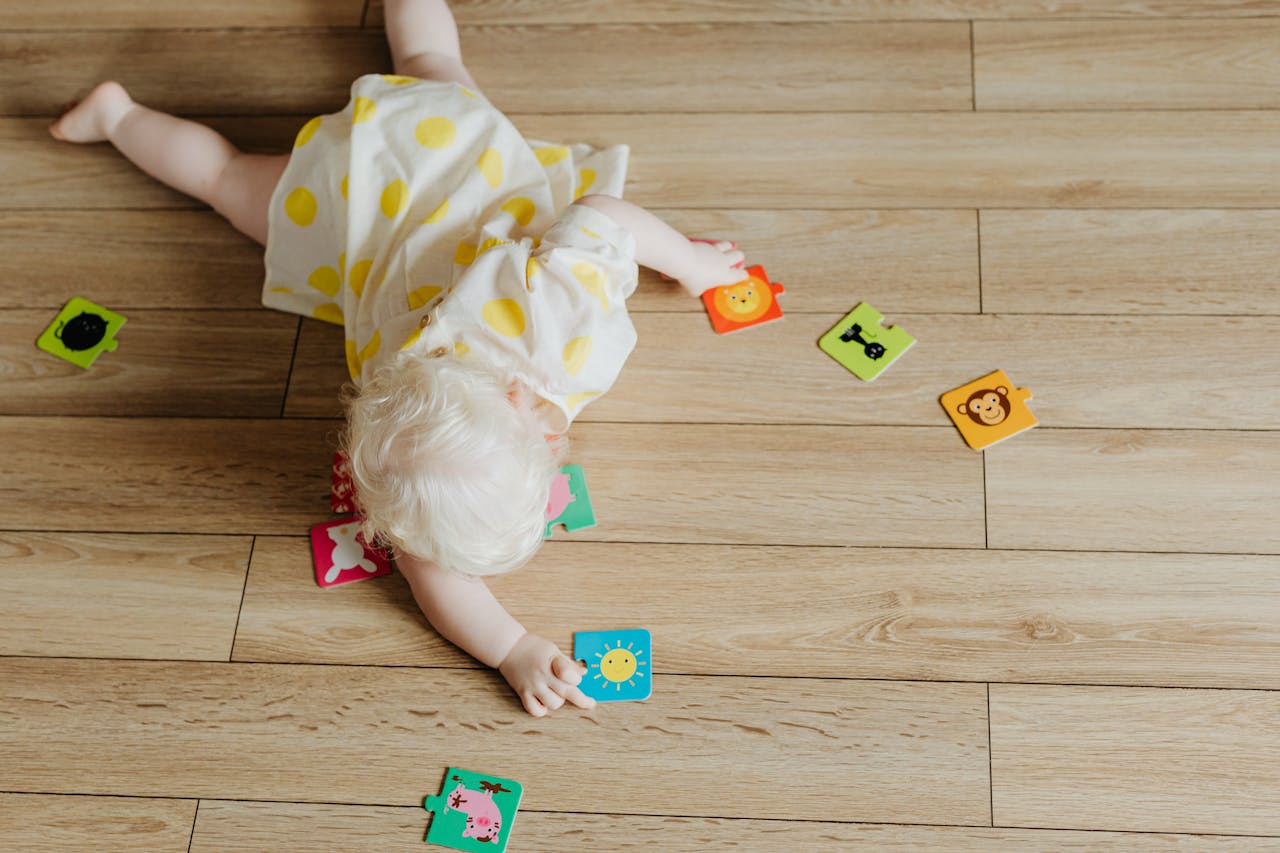 Blond toddler in a white dress with yellow polka dots lying on a wooden floor, reaching for colorful animal-themed puzzle pieces