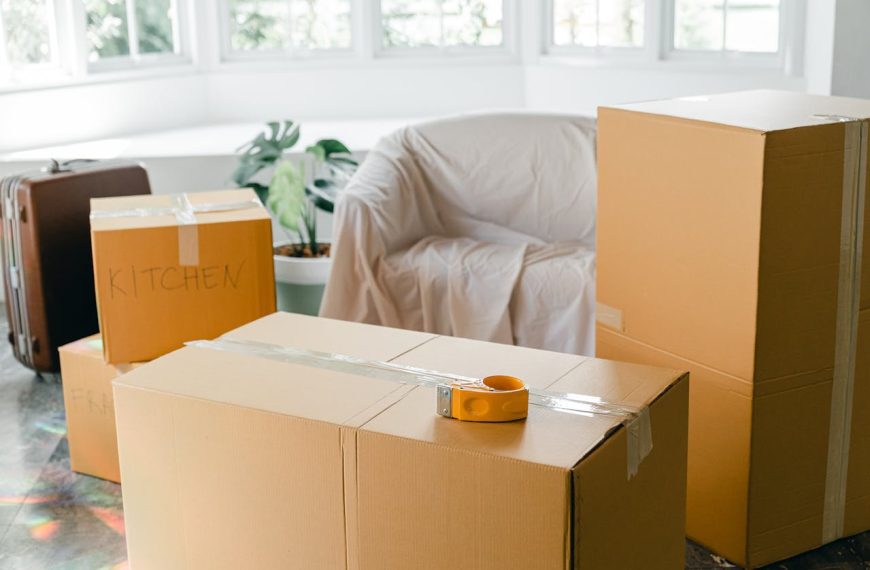 Cardboard moving boxes labeled "KITCHEN" and "FRAGILE", packing tape on top, suitcase and covered armchair in background, indoor plant by window
