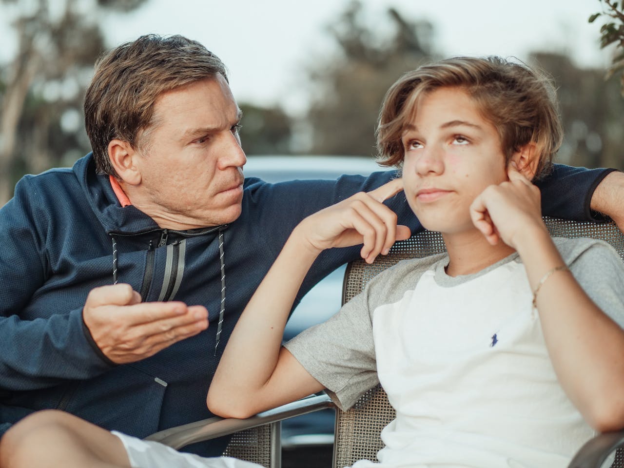 Father looking frustrated while teenage son rolls eyes and covers ears with fingers, sitting outdoors