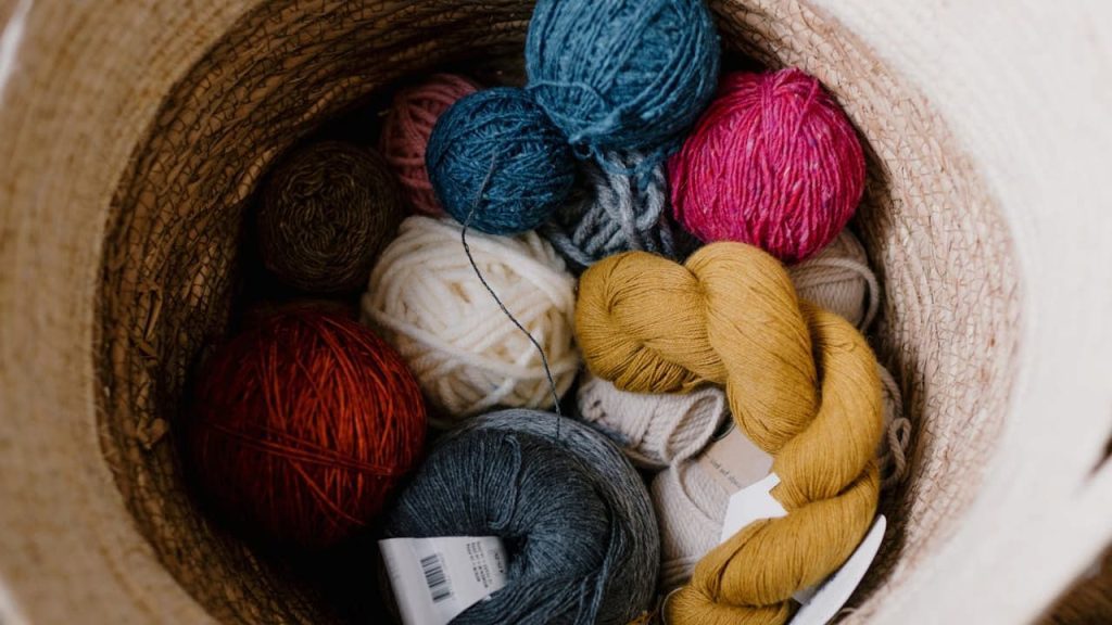 A variety of colorful yarn balls in a basket, including blue, pink, red, white, gray, mustard yellow, and brown, arranged in a circular container