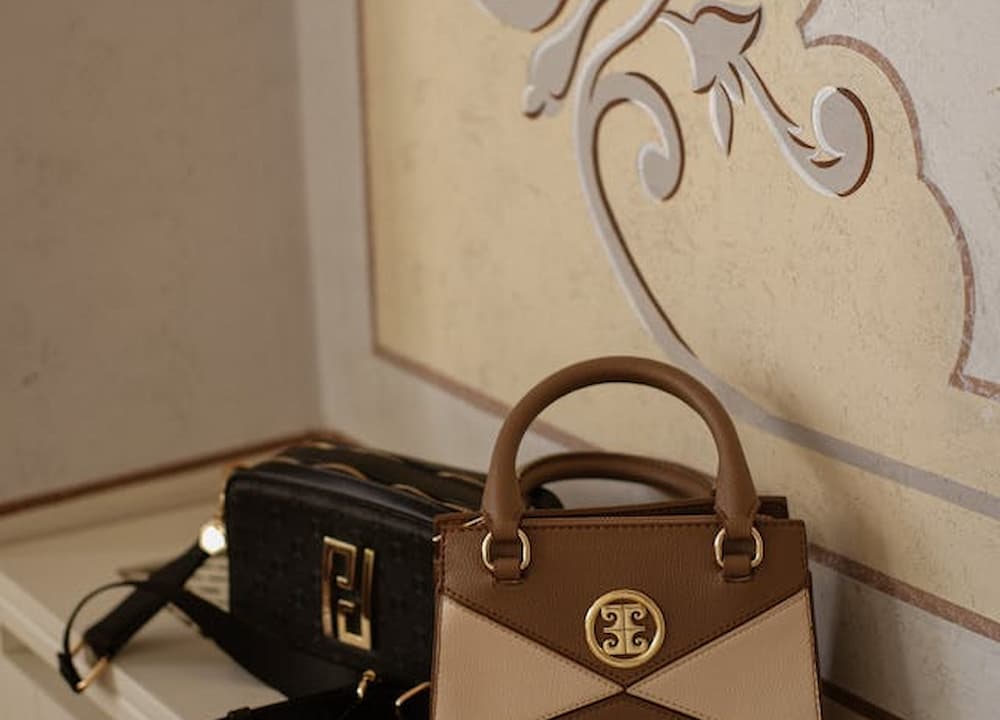 Black Fendi Baguette bag with gold FF logo clasp, resting on a white radiator cover, next to a small two-tone beige and brown top-handle bag with a round gold emblem, decorative wall panel in the background