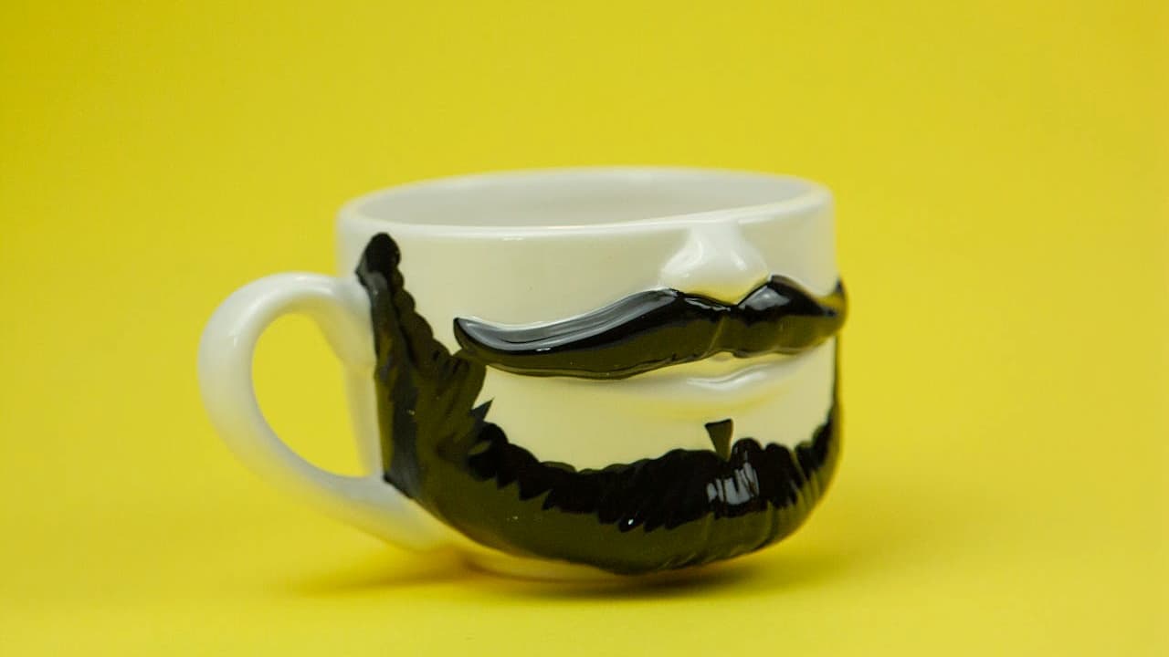 Clear glass mug, painted with black beard and mustache design, yellow background, whimsical and humorous style, novelty drinkware item