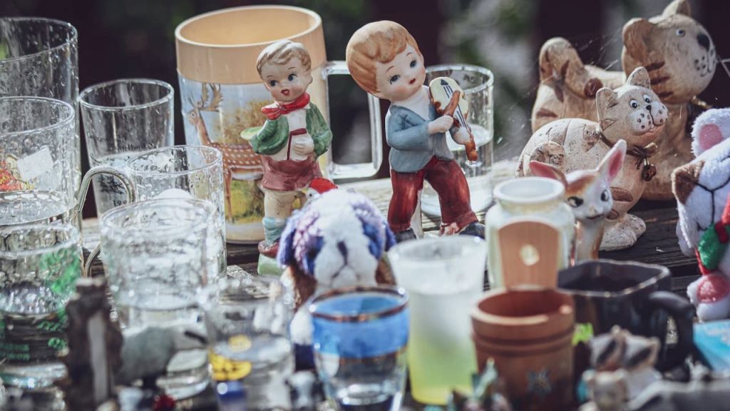Assorted vintage figurines, various glass cups, ceramic decorations, cluttered display on a table, outdoor flea market or yard sale setup, mix of old and collectible items