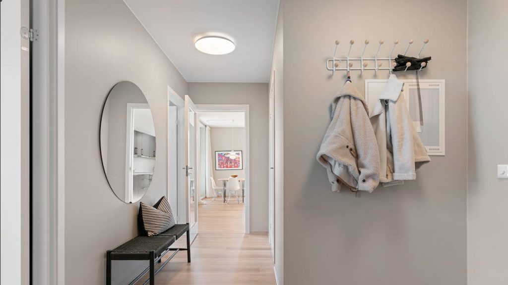 Modern hallway with light wood floors, beige walls, a black bench with a striped pillow, a round wall mirror, two light-colored coats hanging on hooks, and two pairs of shoes neatly placed on the floor