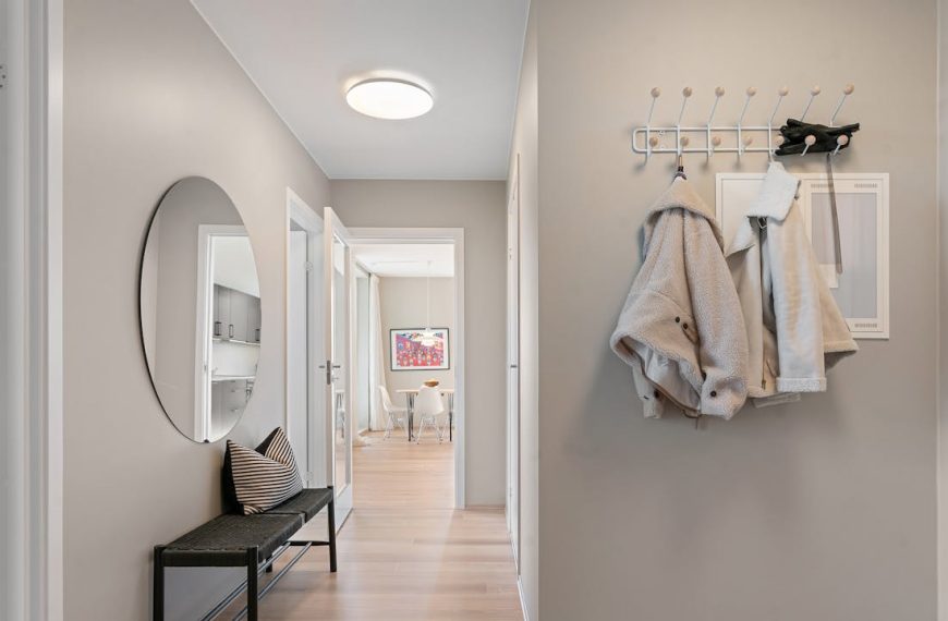 Modern hallway with light wood floors, beige walls, a black bench with a striped pillow, a round wall mirror, two light-colored coats hanging on hooks, and two pairs of shoes neatly placed on the floor