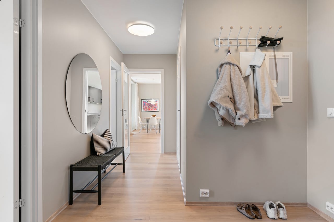 Modern hallway with light wood floors, beige walls, a black bench with a striped pillow, a round wall mirror, two light-colored coats hanging on hooks, and two pairs of shoes neatly placed on the floor