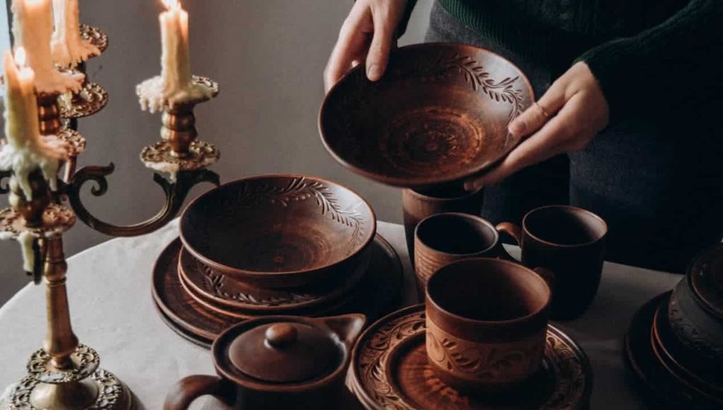 Person holding a carved brown ceramic bowl, surrounded by matching rustic tableware, cups, plates, and a teapot on a white tablecloth, lit by a vintage brass candleholder with burning candles