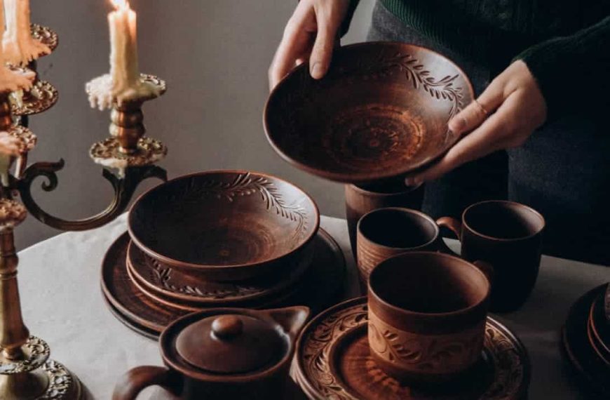 Person holding a carved brown ceramic bowl, surrounded by matching rustic tableware, cups, plates, and a teapot on a white tablecloth, lit by a vintage brass candleholder with burning candles