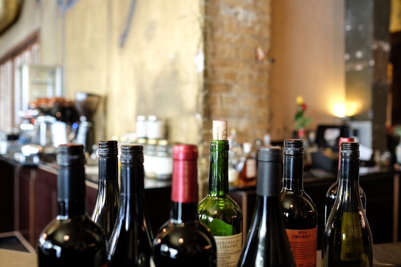 Multiple wine bottles with various labels and closures, placed on a bar counter, blurred café interior in the background, warm lighting, rustic wall texture