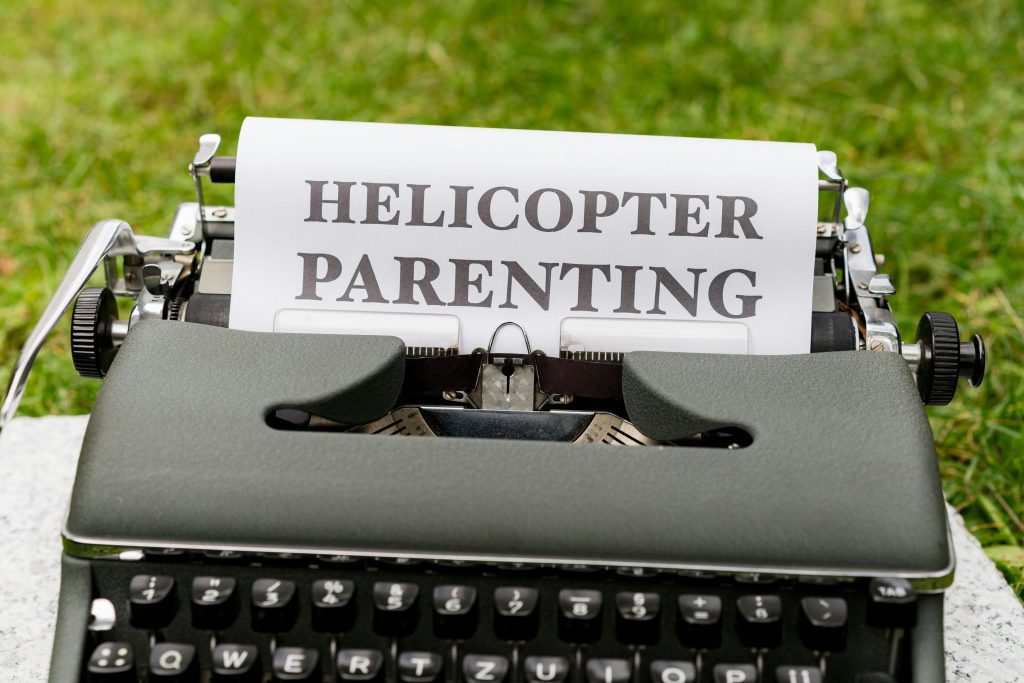 A typewriter with a sheet of paper displaying the bold words “HELICOPTER PARENTING,” set outdoors on green grass