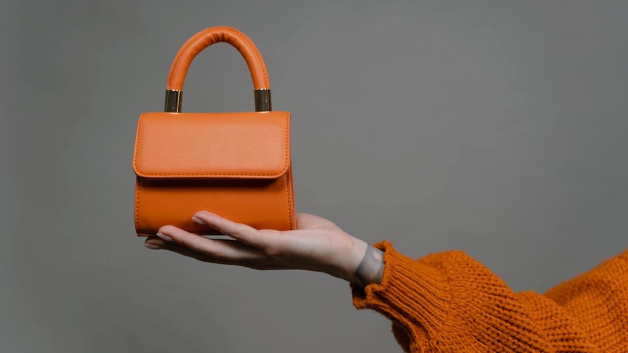  Small orange handbag, structured design, top handle, held by a hand in an orange sleeve, plain gray background