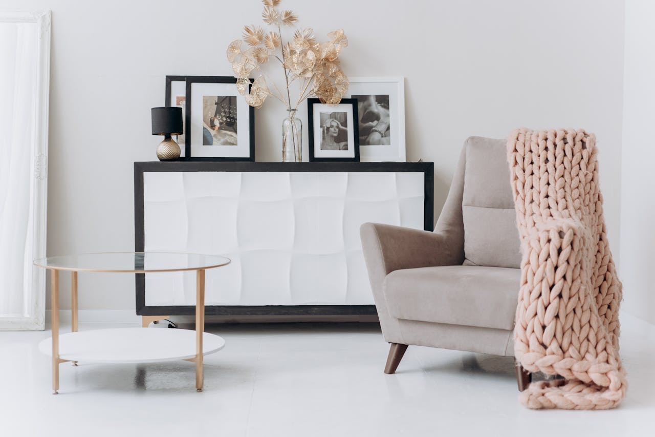 Modern minimalist living room, beige armchair with chunky knit blanket, white sideboard with framed photos, gold and glass coffee table, decorative dried plants in glass vase, Color drenching