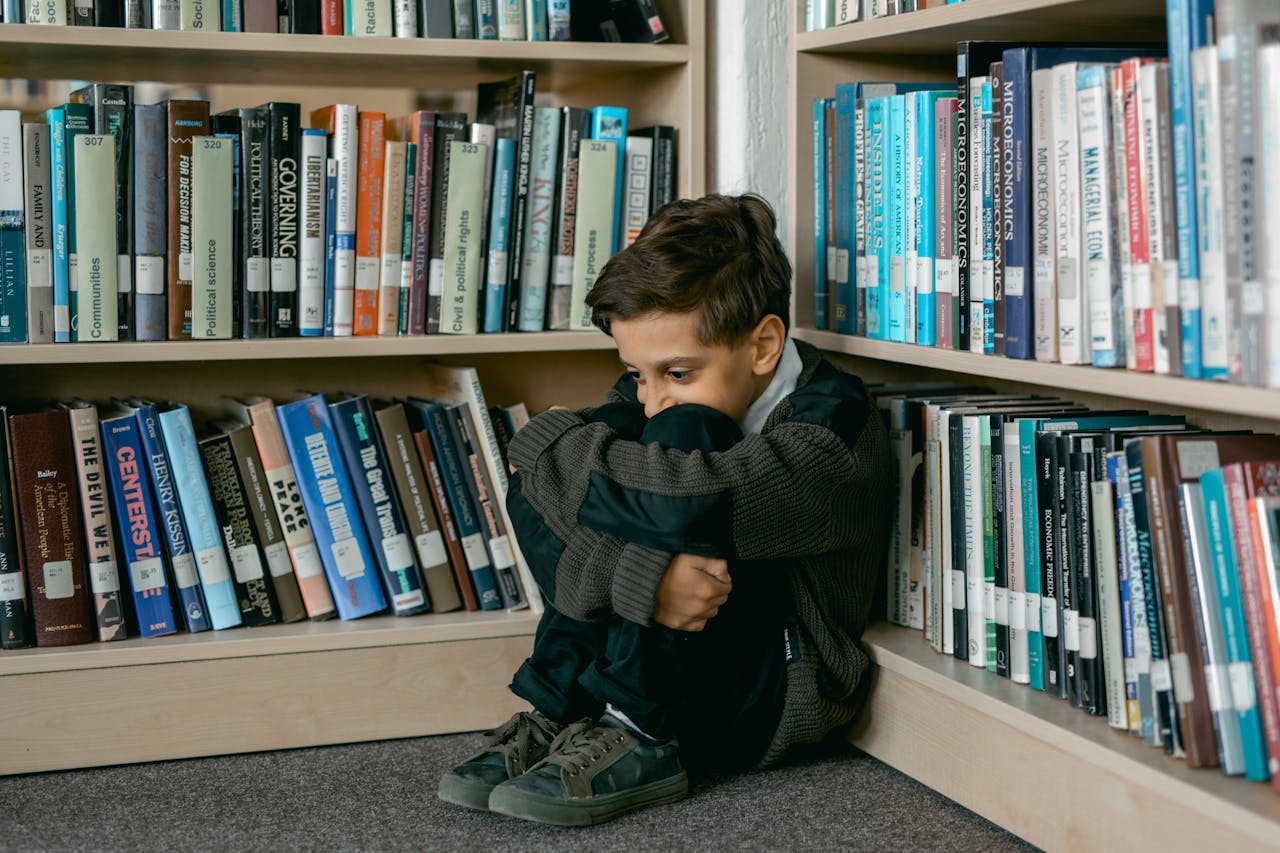 A young boy sits curled up on the floor between bookshelves in a library, hugging his knees, looking down with a pensive expression