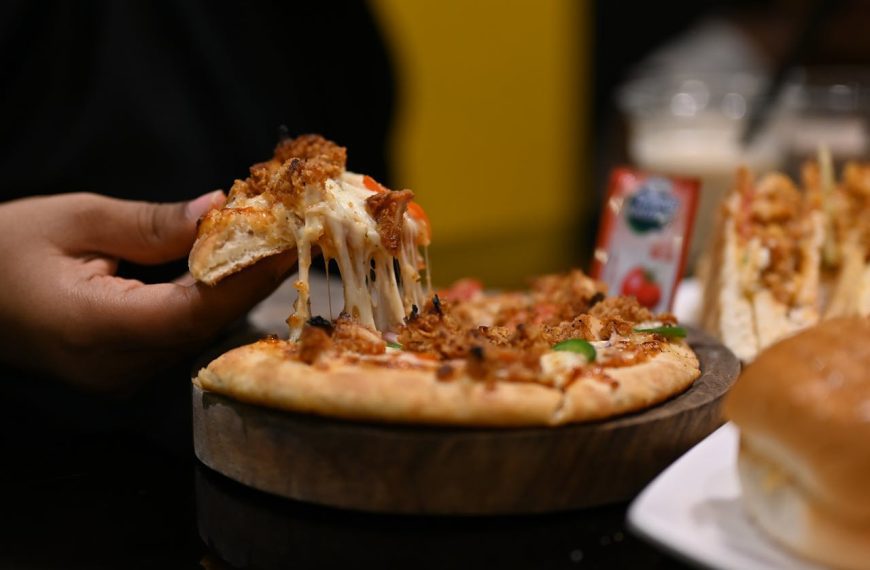 Hand pulling a cheesy slice of pizza topped with meat and vegetables, from a wooden tray, surrounded by sandwiches and condiments