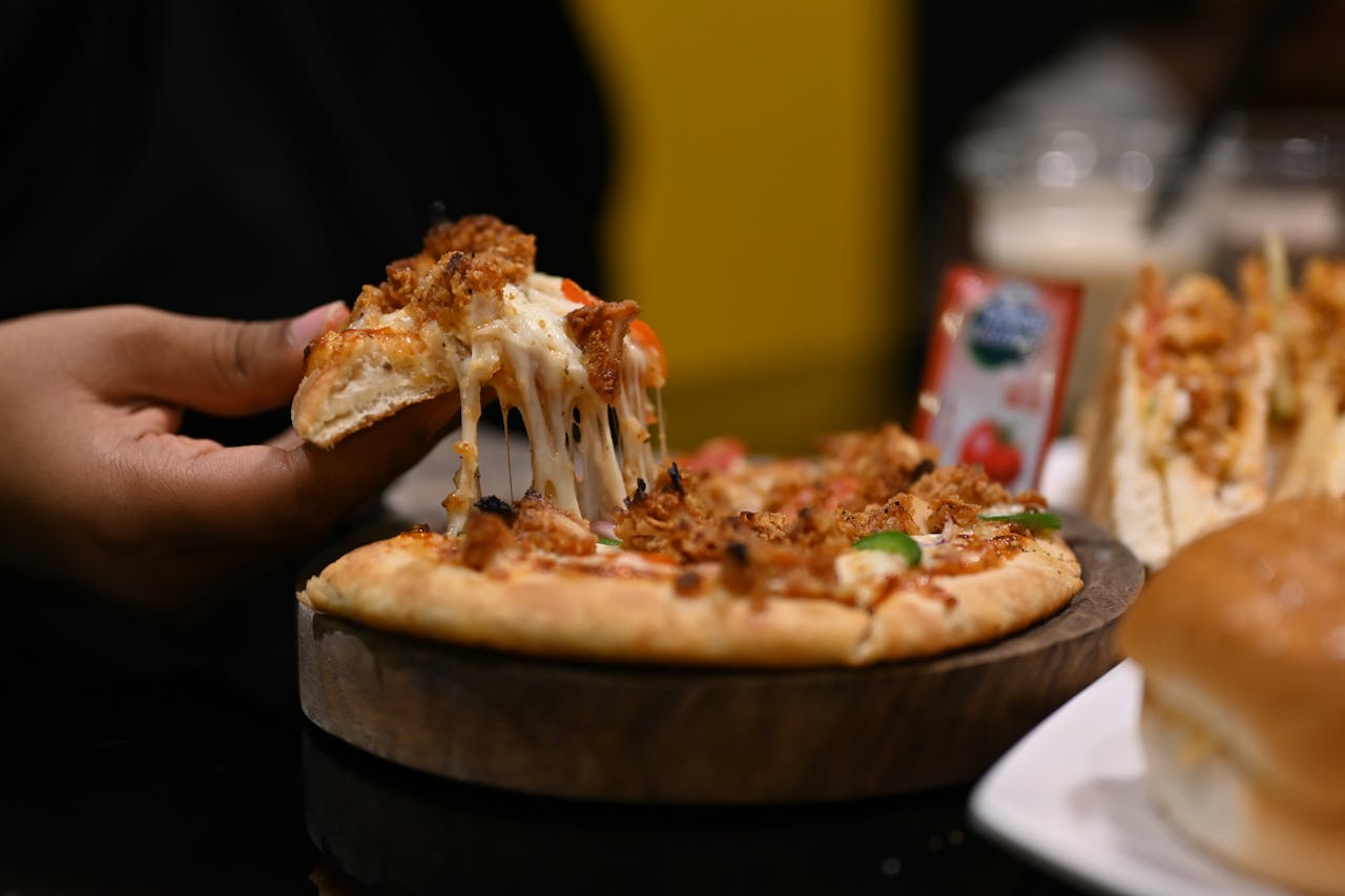 Hand pulling a cheesy slice of pizza topped with meat and vegetables, from a wooden tray, surrounded by sandwiches and condiments