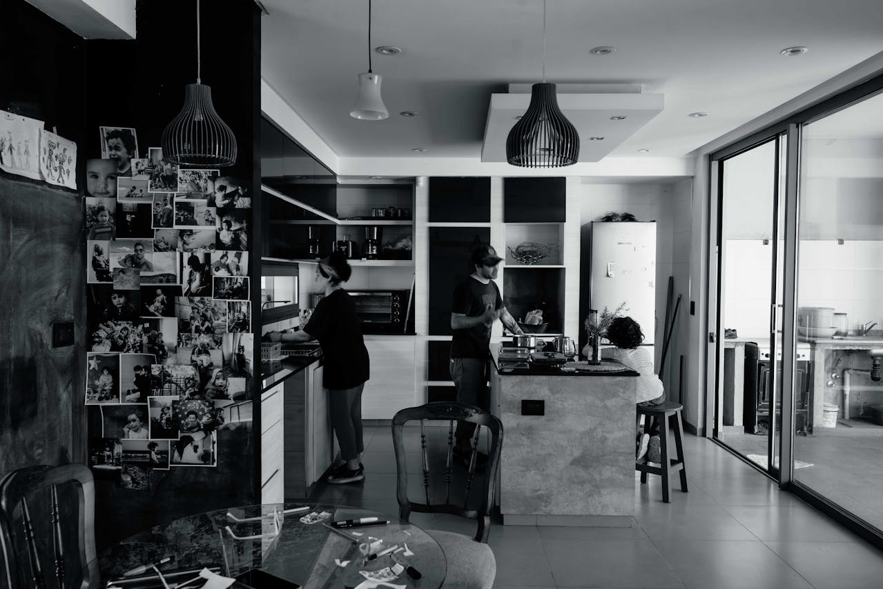 Black and white photo of a modern kitchen with three people, one cooking, one washing dishes, one seated, family photos covering the left wall, glass doors open to a utility area