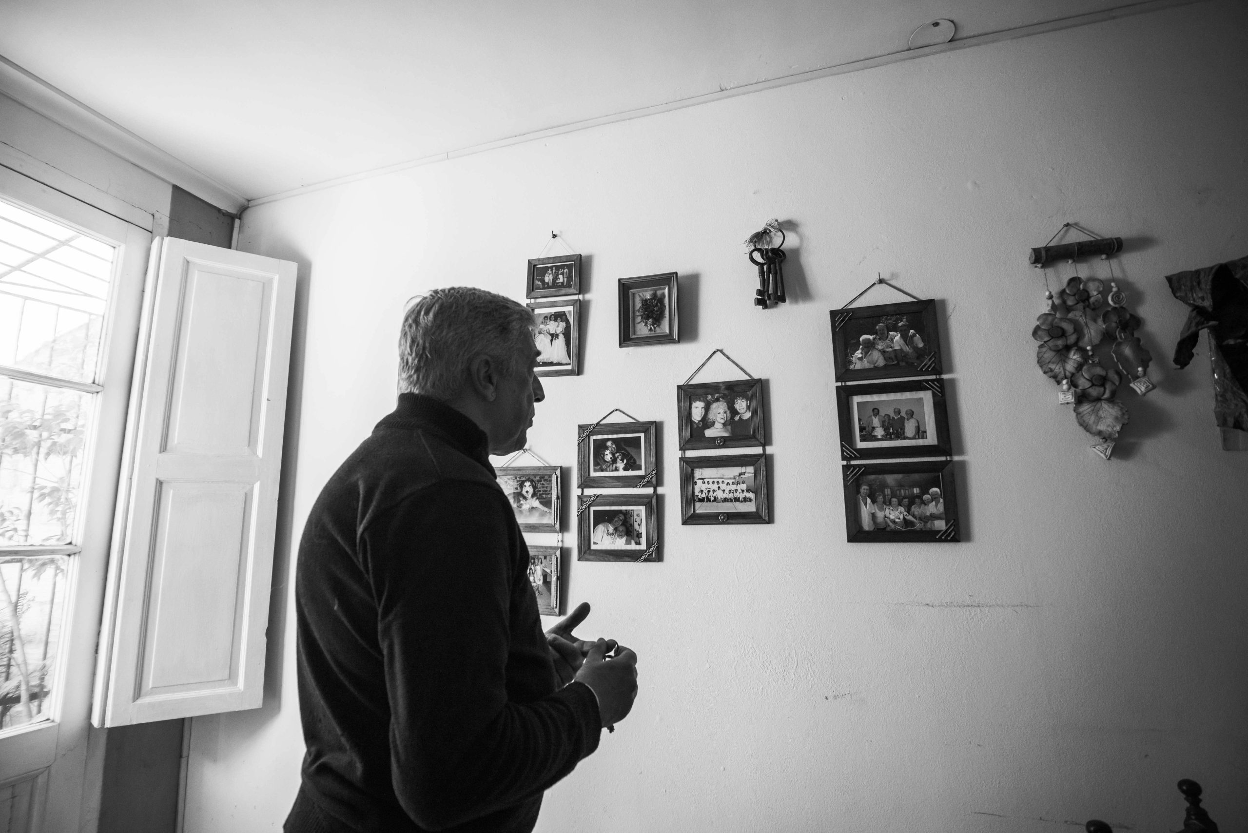 Man standing indoors, looking at a wall filled with framed family photos, natural light coming through an open window, black-and-white image