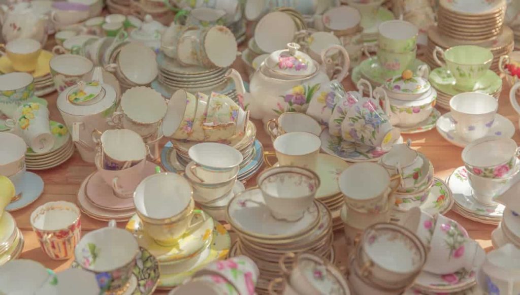 Assorted vintage porcelain teacups and saucers, floral patterns, gold trim, pastel colors, stacked closely on a wooden table, sunlit setting