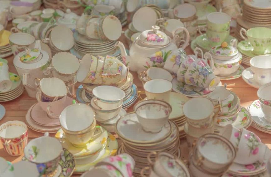 Assorted vintage porcelain teacups and saucers, floral patterns, gold trim, pastel colors, stacked closely on a wooden table, sunlit setting
