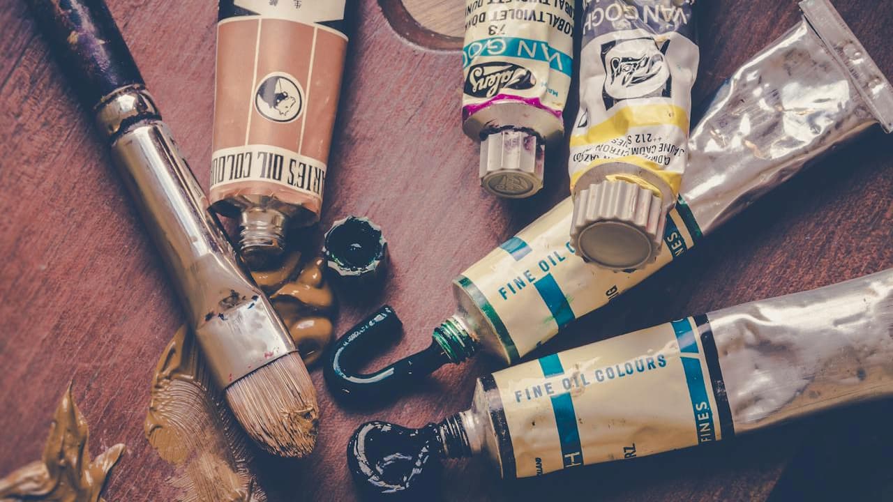 Used paintbrush and squeezed paint tubes on a wooden surface, worn labels, visible smudges of paint, earthy and pastel color tones, artistic workspace setting