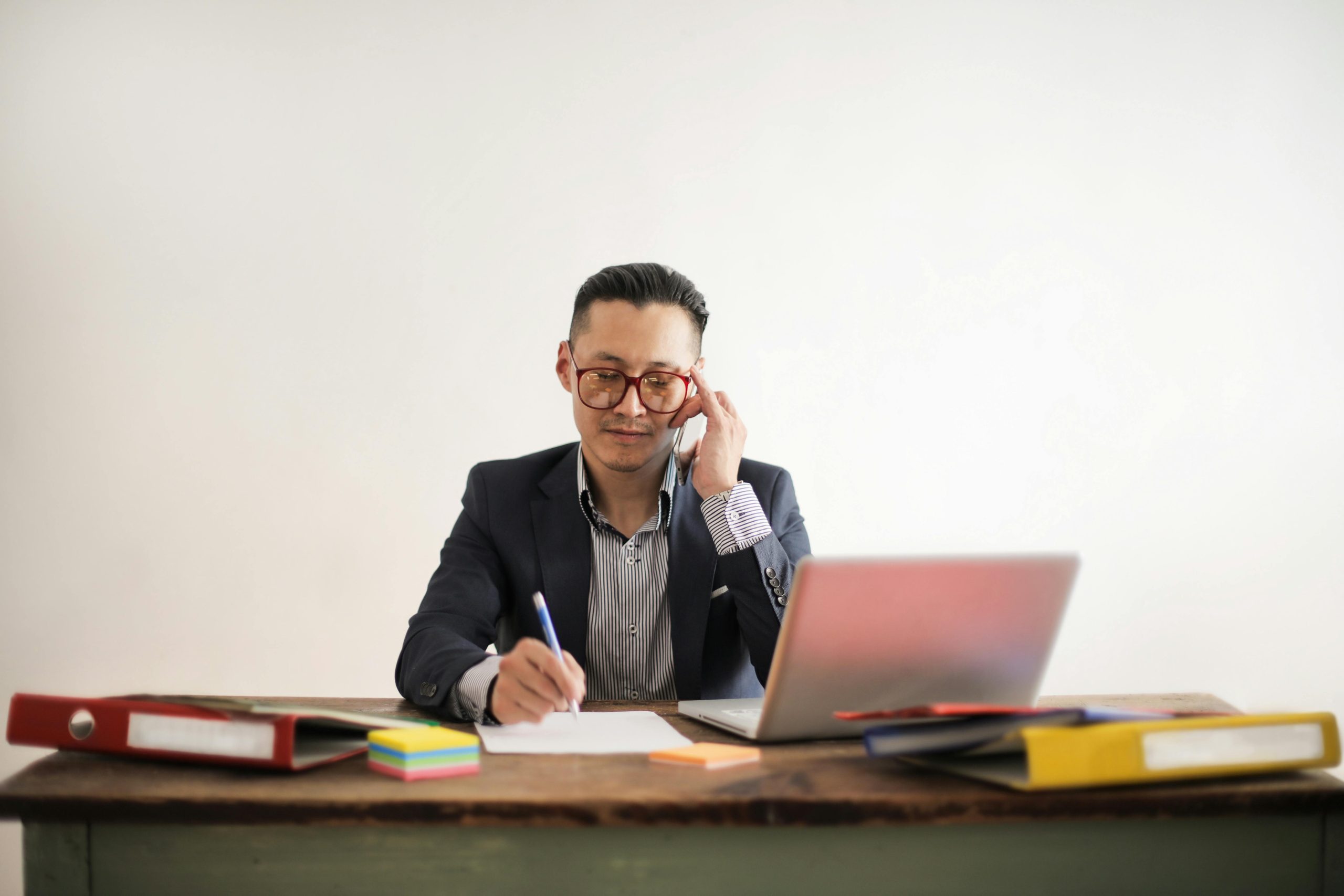 Man in a suit sitting at a wooden desk, talking on the phone, writing on paper with a pen, using a laptop, surrounded by colorful folders and sticky notes, focused expression, plain white background
