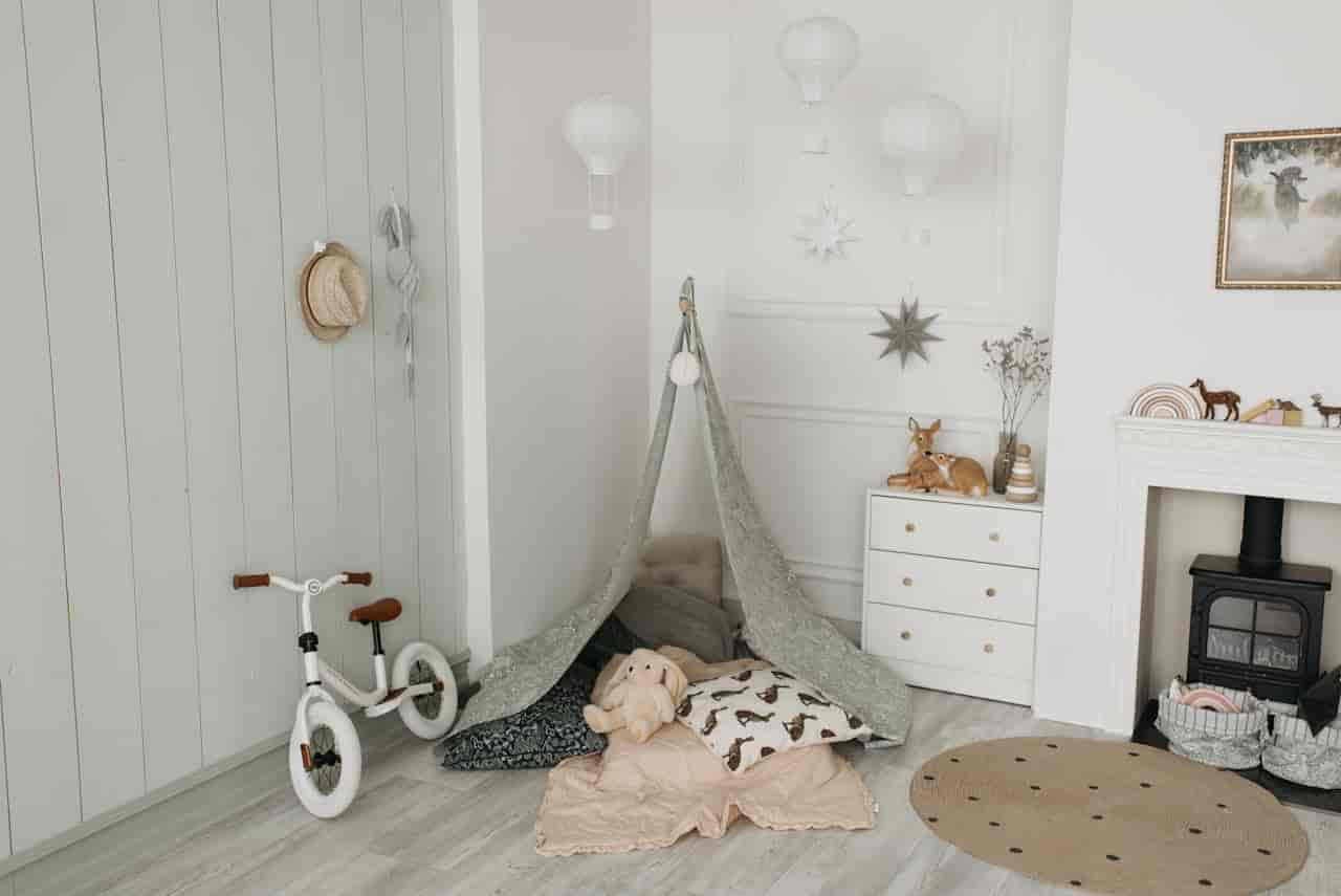 Scandinavian-style children's playroom, indoor teepee tent with pillows and stuffed animals, white balance bike, dresser with toys, soft rug, neutral decor with star wall hangings and hot air balloon lights