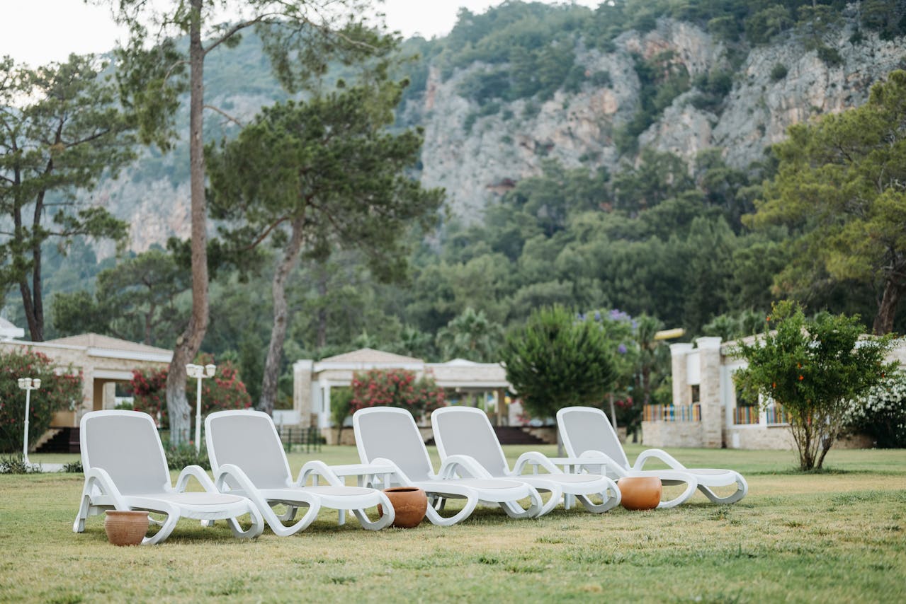 Six white lounge chairs, arranged on a grassy lawn, small terracotta stools beside them, with trees, bushes, and cottages in the background, mountains rising behind