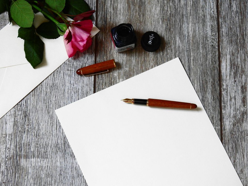 Blank white paper with a wooden fountain pen resting on it, open ink bottle nearby, rose flower and cream-colored envelopes on a rustic wooden surface, scene suggests letter writing or creative expression