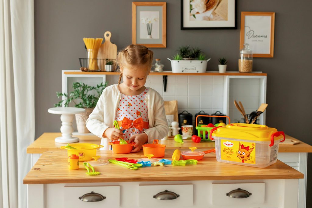 Child playing with colorful plastic building blocks, wooden table surface, yellow storage bin, white toy shelf in background, framed pictures and plants on wall, cozy and organized indoor play area
