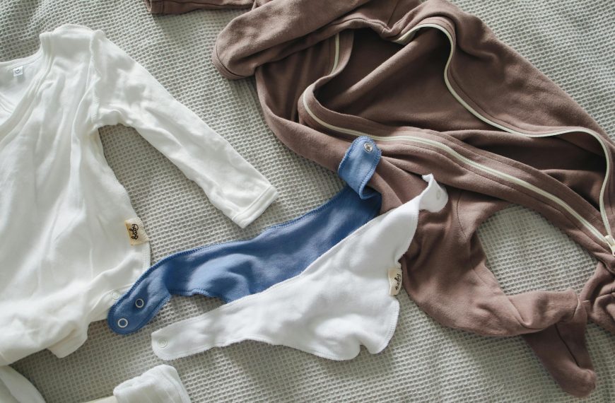Baby clothes laid out on a textured blanket, including a white long-sleeve onesie, a brown zip-up suit, and two soft bibs in white and blue, all made of cozy fabric