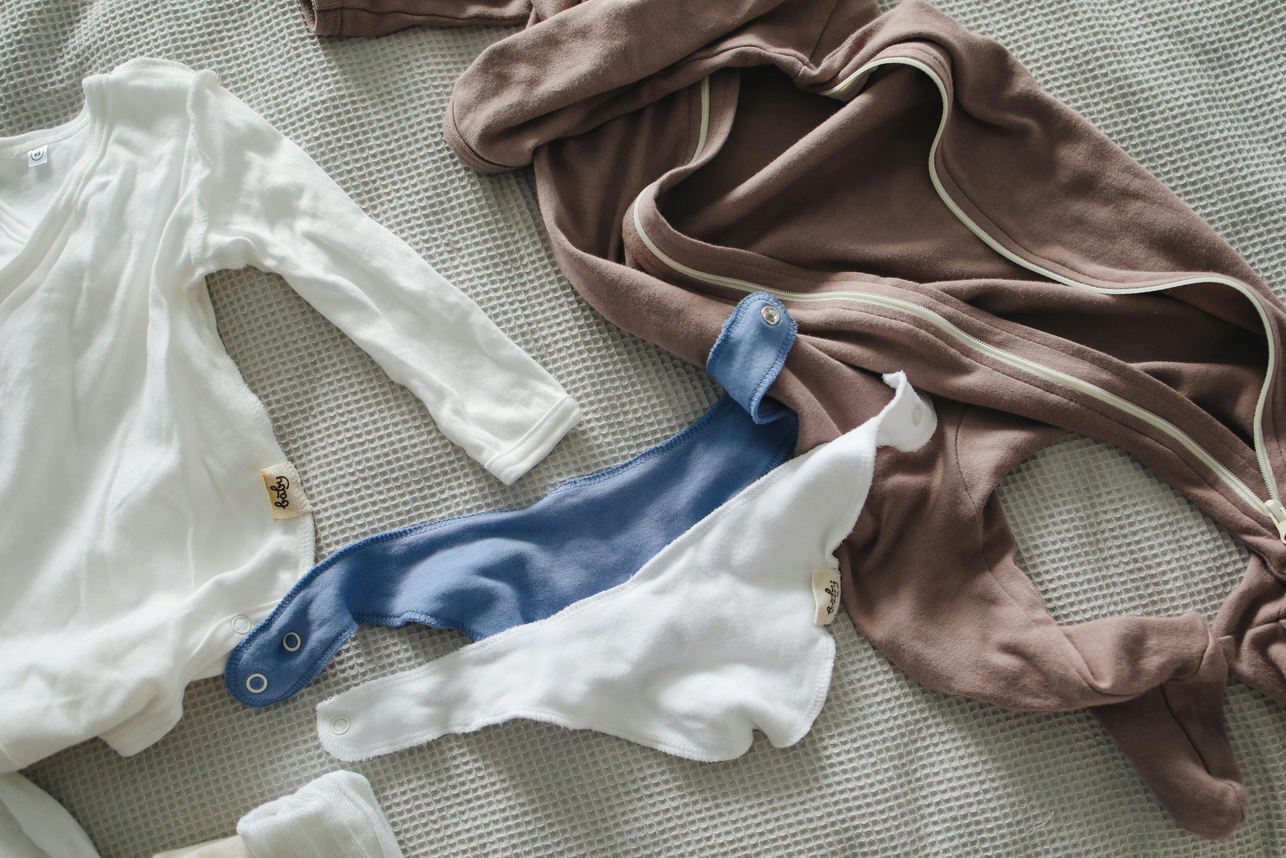 Baby clothes laid out on a textured blanket, including a white long-sleeve onesie, a brown zip-up suit, and two soft bibs in white and blue, all made of cozy fabric